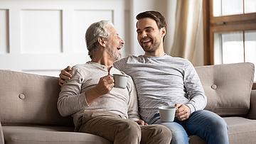 Ein Rentner und ein Mann sitzen lachend auf dem Sofa.