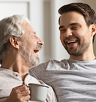Ein älterer Herr sitzt lachend mit einer Tasse in der Hand auf dem Sofa. Ein jünger Mann sitzt daneben und hat den Arm um den Rentner gelegt.