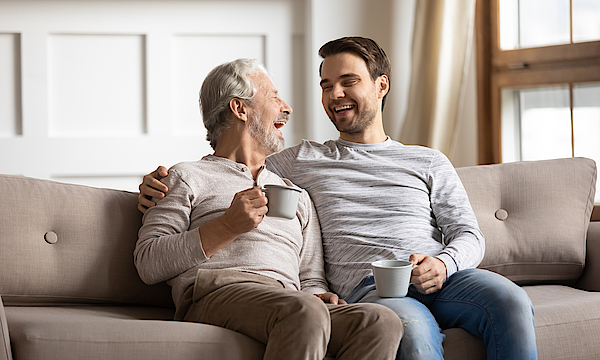 Ein älterer Herr sitzt lachend mit einer Tasse in der Hand auf dem Sofa. Ein jünger Mann sitzt daneben und hat den Arm um den Rentner gelegt.