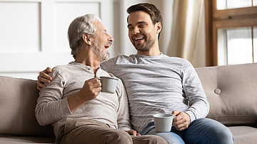 Ein Rentner und ein Mann sitzen lachend auf dem Sofa.
