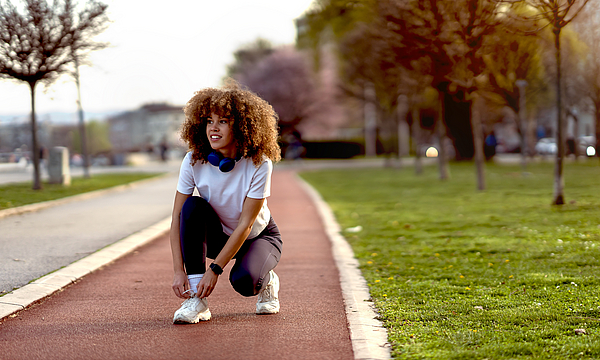 Junge Frau, die bei Sonnenuntergang auf einer Laufbahn in einem Stadtpark Schuhe bindet. Sie trägt Sportkleidung und trainiert offensichtlich, vielleicht im Zone-2-Training.