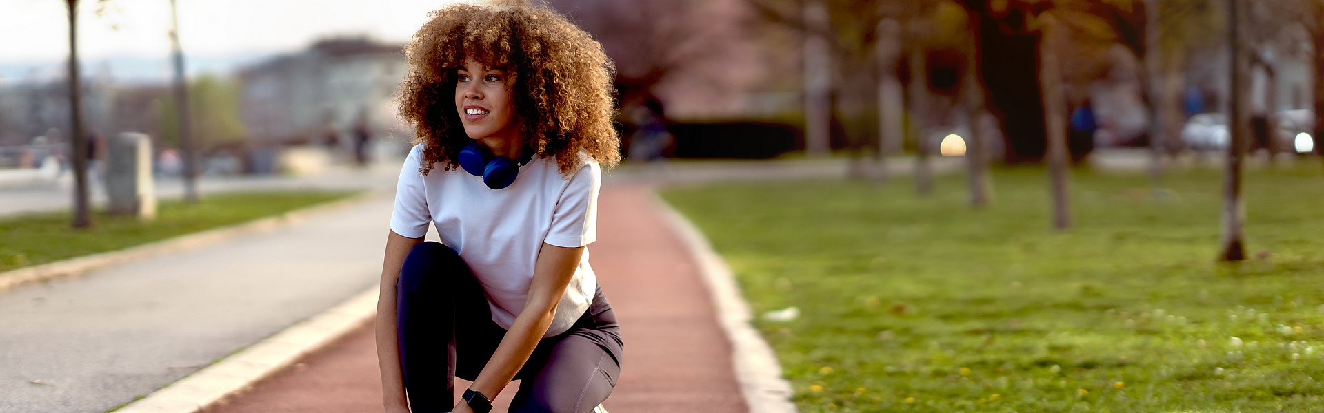 Junge Frau, die bei Sonnenuntergang auf einer Laufbahn in einem Stadtpark Schuhe bindet. Sie trägt Sportkleidung und trainiert offensichtlich, vielleicht im Zone-2-Training.