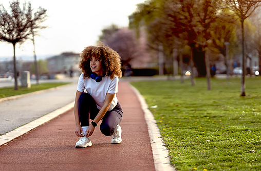 Junge Frau, die bei Sonnenuntergang auf einer Laufbahn in einem Stadtpark Schuhe bindet. Sie trägt Sportkleidung und trainiert offensichtlich, vielleicht im Zone-2-Training.