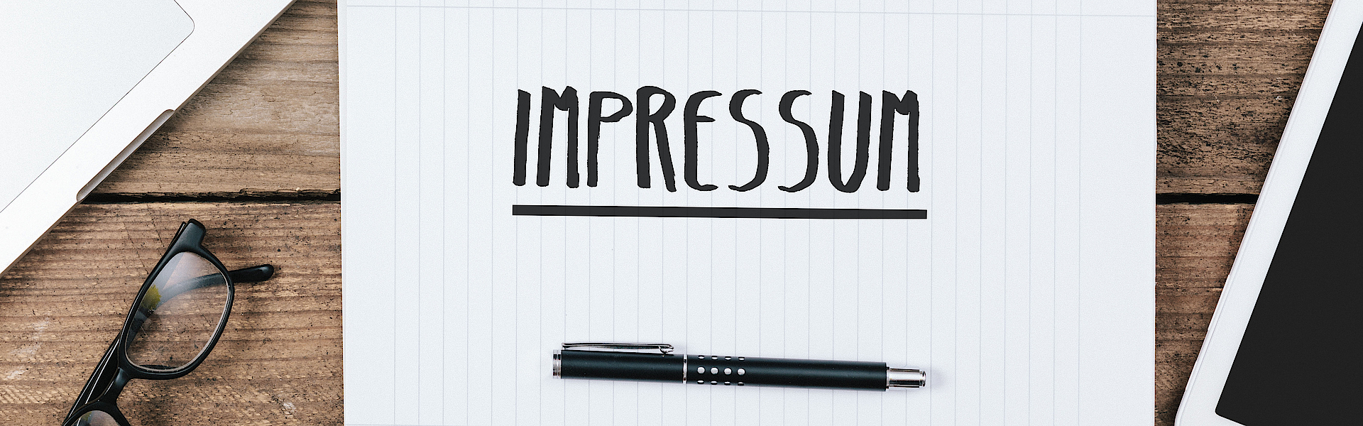 Auf einem Schreibtisch liegt ein Blatt Papier. Darauf ist mit schwarzem Stift das Wort Impressum geschrieben. 