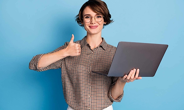 Eine Frau hält einen Laptop in der Hand. Sie hält den rechten Daumen nach oben und freut sich.