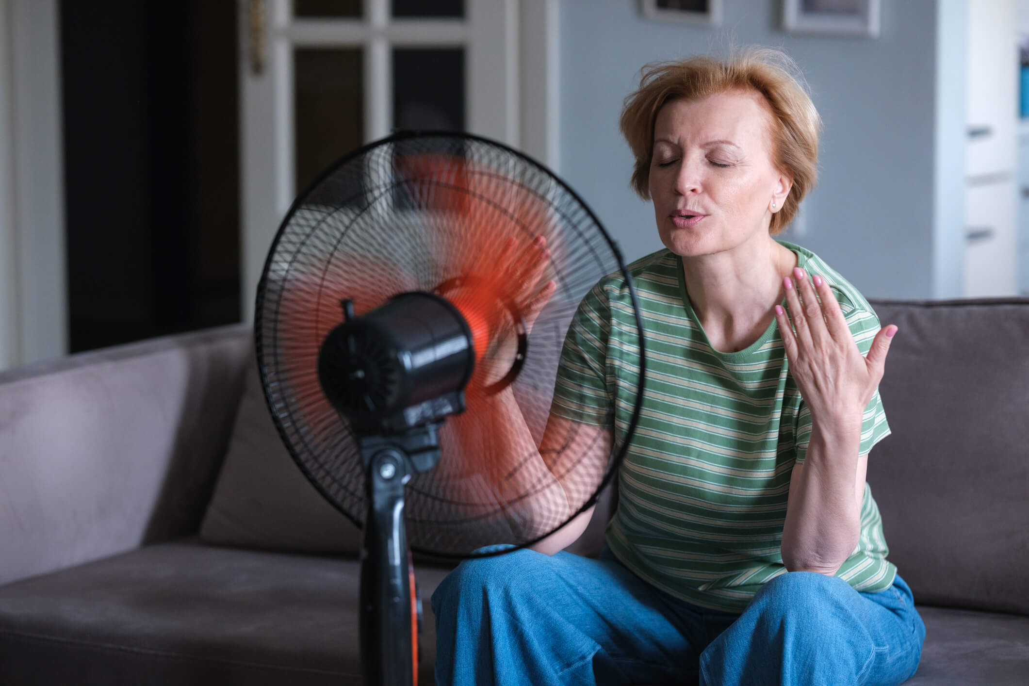 Frau im mittleren Altern sitzt vor einem Ventilator, ihr ist heiß.