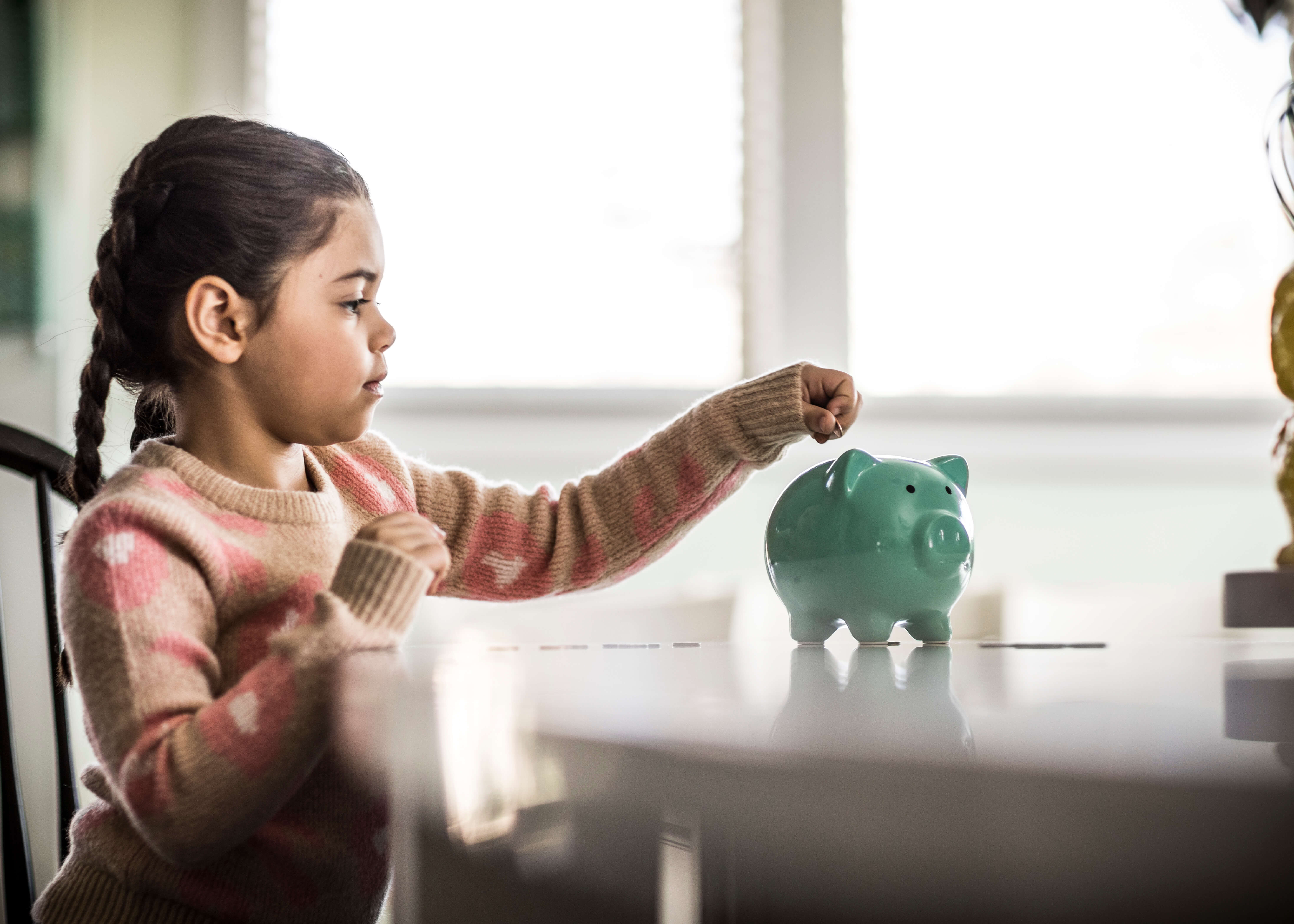 Kleines Mädchen steht am Tisch und wirft Geld in ein grünes Sparschwein